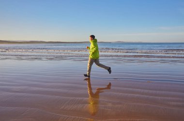 Yeşil ceketli enerjik genç adam Essaouira, Fas 'ta okyanus boyunca koşuyor. Manzaralı kıyı şeridinin tadını çıkarıyor, aktif bir yaşam tarzını kucaklıyor.