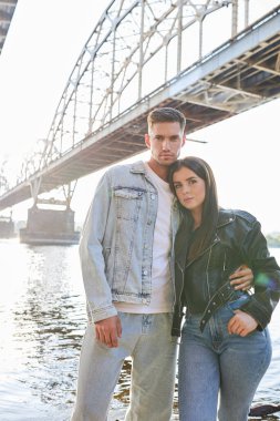 Arka planda büyük bir köprüyle dışarıda poz veren genç ve şık bir çift..