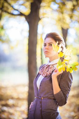 Sonbahar ormanının büyüleyici güzelliği arasında yakalanmış zarif esmer bir kız zarafet ve sükunetle ışıldıyor. Düşen yaprakların canlı renkleri ve benekli güneş ışığı onun varlığı için büyüleyici bir zemin oluşturur..