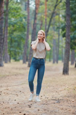Çam ormanında poz veren doğal makyajlı sarışın kadın. Güzel mavi gözlü, bej tişörtlü beyaz kadın yaz tatilinin tadını çıkarıyor. Vahşi yaşam ve insanlar kavramı.
