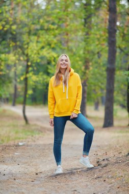 Çekici mavi gözlü sarışın kadın şehir parkında yürüyor. Sarı kapüşonlu, pembe çantalı, mutlu görünen ve gülümseyen bir kız. Sonbahar parkında eğlenen neşeli genç bir kadının portresi. Doğada rahatla..