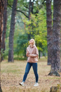 Çekici mavi gözlü sarışın kadın şehir parkında yürüyor. Kız bej kapüşonlu, pembe bir çanta giyiyor ve mutlu görünüyor ve gülümsüyor. Sonbahar parkında eğlenen neşeli genç bir kadının portresi. Doğada rahatla..