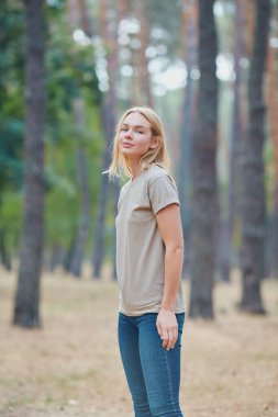 Çam ormanında poz veren doğal makyajlı sarışın kadın. Güzel mavi gözlü, bej tişörtlü beyaz kadın yaz tatilinin tadını çıkarıyor. Vahşi yaşam ve insanlar kavramı.