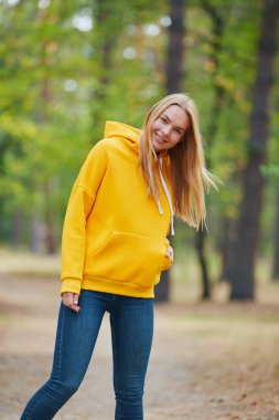 Sarı kapüşonlu mavi gözlü sarışın çam ormanında yürüyor. Sonbahar parkında eğlenen neşeli genç bir kadının portresi. Doğada rahatla..