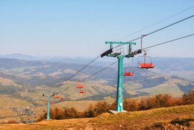 Bu büyüleyici fotoğraf, güneşli bir günün sıcacık ışıltısında güneşlenen resimli sonbahar dağlarının arka planına yerleşmiş eski bir teleferiği gösteriyor. Nostalji ve engebeli bir güzellik, doğa ve insan mühendisliği hissi yayar..