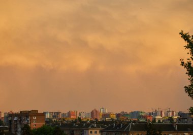 A soft pastel sunset with light pink clouds floating above silhouetted rooftops and distant city buildings. The warm evening sky transitions from yellow to light blue, creating a peaceful urban scene. clipart