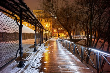 Parkta kış akşamı, karla kaplı banklar, parlak ışıklar beyaz karı aydınlatıyor, yeni yıl arifesinde kar yağıyor..