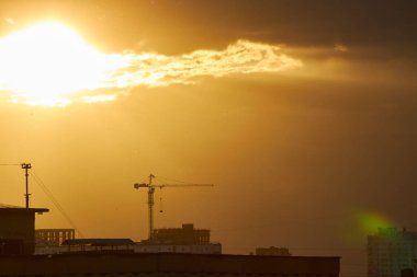 A bright golden sunset casts a warm glow over silhouetted buildings and a construction crane. The intense light filters through clouds, creating a dramatic and radiant urban scene clipart