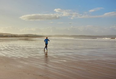 Genç bir kadın, Fas 'ın Essaouira sahilinde serinletici bir koşunun keyfini çıkarırken, dalgalar kıyı zeminine hafifçe vuruyor.