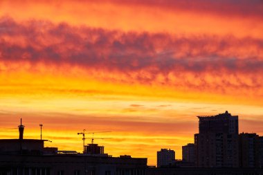 Yoğun turuncu ve sarı renkli canlı bir günbatımı, binaların siluetlerini ve bir inşaat vincini ön plana çıkarıyor. Bulutlu bir gökyüzünün altında dramatik bir ufuk çizgisi oluşturuyor..