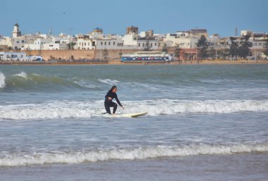 Genç bir dişi sörfçü, sörf kültürünün manzaralı sahil şeridine karşı heyecanını ve güzelliğini yakalayan Essaouira, Fas kıyıları boyunca sörf tahtasında zarif bir şekilde sörf yapıyor..