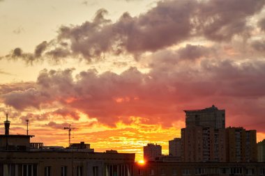 Canlı bir günbatımı, koyu bulutlarla çelişen sıcak turuncu ve kırmızı renklerle gökyüzünü boyar. Şehrin ufuk çizgisi siluetlidir, bir vinç ve binalar dramatik manzaraya kentsel bir dokunuş katar..