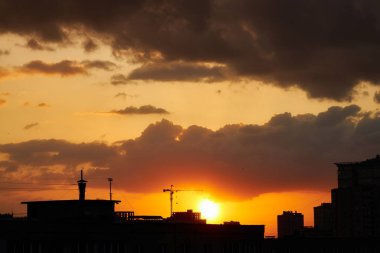Yoğun turuncu ve sarı renkli canlı bir günbatımı, binaların siluetlerini ve bir inşaat vincini ön plana çıkarıyor. Bulutlu bir gökyüzünün altında dramatik bir ufuk çizgisi oluşturuyor..