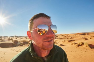 Güneş gözlüğü takan bir adamın yakın plan portresi. Bir grup turist gözlüklerin yansımasından görülebilir. Sahra Çölü, Fas 'ta.