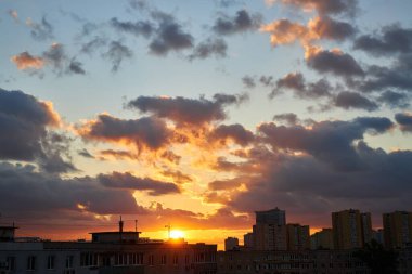 Çarpıcı bir günbatımı gökyüzünü turuncu, sarı ve morun canlı tonlarıyla doldurur. Silüetli çatılar ve binalar, ateşli bulutlarla tezat oluşturarak dramatik ve renkli bir şehir manzarası yaratıyor..