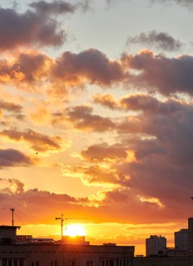 Çarpıcı bir günbatımı gökyüzünü turuncu, sarı ve morun canlı tonlarıyla doldurur. Silüetli çatılar ve binalar, ateşli bulutlarla tezat oluşturarak dramatik ve renkli bir şehir manzarası yaratıyor..