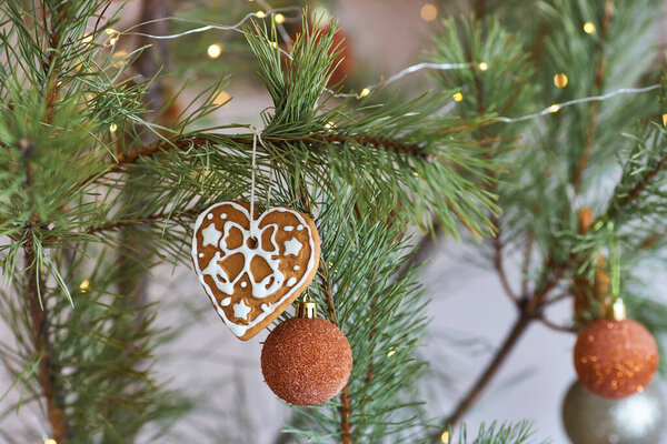 Close-up of a heart-shaped gingerbread cookie with white icing and a glittery orange bauble hanging on a pine branch. Warm festive lights add to the cozy holiday atmosphere.