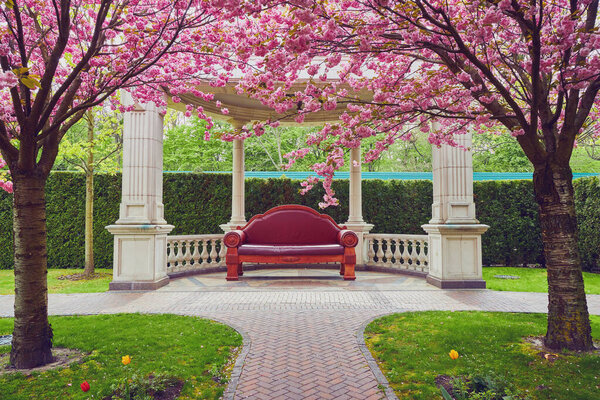 A spring day in the Sakura Park, featuring a grand leather sofa under a gazebo, creating an elegant and comfortable retreat amid blooming cherry blossoms.