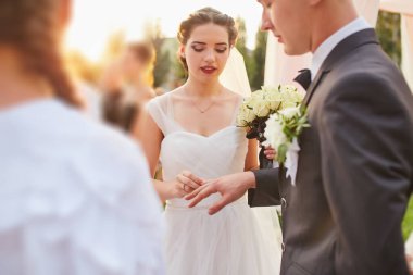 Damat tören sırasında gelinin parmağına bir alyans takar. Gelin elinde bir buket beyaz gül tutarken ikisi de süslü bir kemerin altında akan kumaşla yemin ediyor..