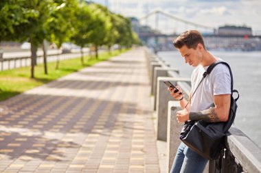 Beyaz tişört ve kot pantolon giyen, elinde akıllı telefon tutan neşeli bir genç. Siyah bir sırt çantası, yeşil ağaçlar ve arka planda bir köprüyle nehir kenarındaki bir geçitte duruyor..