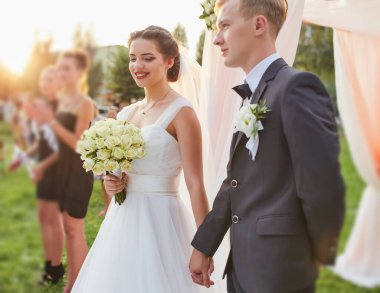 Beyaz elbiseli bir gelin elinde bir gül buketi ve siyah takım elbiseli bir damat çiçeklerle süslenmiş bir düğün kemerinin altında durup, açık hava seremonileri sırasında gülümsüyor ve el ele tutuşuyorlar..