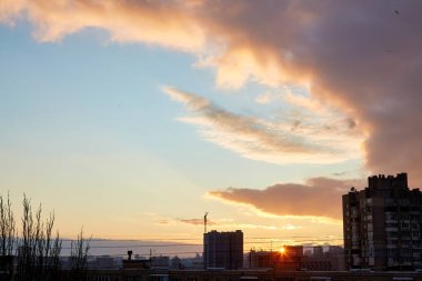 Şehir silueti üzerinde hareketli bulutlarla dramatik bir günbatımı. Ufuk batan güneşle parlıyor, şehir manzarasına ve siluet binalarına altın bir ışık tutuyor..