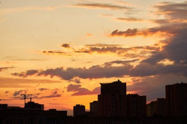 Canlı bir günbatımı, koyu bulutlarla çelişen sıcak turuncu ve kırmızı renklerle gökyüzünü boyar. Şehrin ufuk çizgisi siluetlidir, bir vinç ve binalar dramatik manzaraya kentsel bir dokunuş katar..