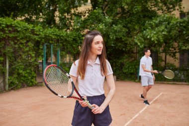 Genç bir bayan tenisçi raketini güvenli bir şekilde tutarken, takım arkadaşı arka planda hazırlanıyor. Yeşillik, açık hava atmosferini güçlendiriyor..