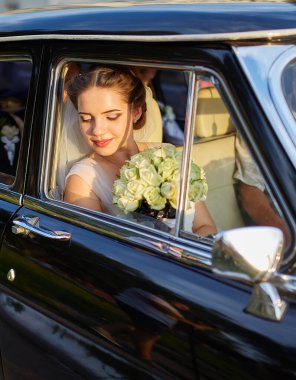 Siyah bir arabanın içinde oturan gelin elinde bir buket beyaz gülle pencereden dışarı dikkatlice bakıyor. Gün batımında yakalanmış, sakin ve zarif gelinliğini vurguluyor..