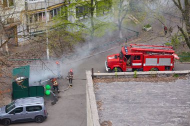 İtfaiyecilerden oluşan bir ekip yerleşim yerinin yakınındaki yangını söndürüyor. Yakınlarda kırmızı bir itfaiye kamyonu var ve sakinleri acil durum tepkisini izliyor. Yanan bölgeden duman yükseliyor..