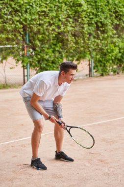 Odaklanmış genç bir adam kil tenis kortunda servis yapmaya hazırlanıyor. Beyaz polo tişört ve gri şort giymiş, elinde bir tenis raketi tutmakta ve harekete geçmeye hazırdır..
