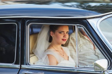 Beyaz bir duvak takan, şık bir elbise giyen, klasik siyah bir arabanın içinde oturan güzel bir gelin. Güneş ışığı, pencereden bakarken onun huzurlu ve neşeli yüzünü hafifçe aydınlatıyor..