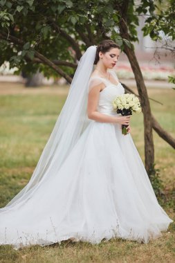Uzun peçeli, beyaz elbiseli neşeli bir gelin ve bir buket beyaz gül dışarıda, yemyeşil bir bahçede duruyor. Özel gününde ışıl ışıl ve zarif görünüyor..