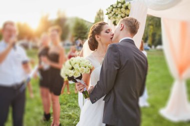 Yeni evliler düğün törenlerinde romantik bir öpücük paylaşıyorlar. Gelin, misafirlerle çevrili bir buket beyaz gül ve yumuşak kumaş ve çiçeklerle süslenmiş bir kemer tutuyor..