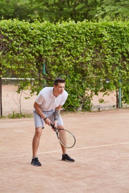 Odaklanmış genç bir adam kil tenis kortunda servis yapmaya hazırlanıyor. Beyaz polo tişört ve gri şort giymiş, elinde bir tenis raketi tutmakta ve harekete geçmeye hazırdır..