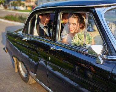 Beyaz bir duvak takan, şık bir elbise giyen, klasik siyah bir arabanın içinde oturan güzel bir gelin. Güneş ışığı, pencereden bakarken onun huzurlu ve neşeli yüzünü hafifçe aydınlatıyor..