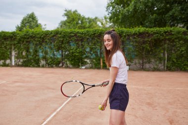 Genç bir kadın kil tenis kortunda hizmet etmeye hazırlanıyor. Elinde beyaz polo ve donanma şortu giymiş bir tenis topu ve raket tutuyor..