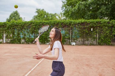 Spor kıyafetli genç bir kadın açık hava kil kortunda enerjik bir şekilde tenis oynuyor, salıncağın ortasında yakalandı, arka planda yemyeşil bir şey var..