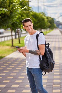 Beyaz tişört ve kot pantolon giyen, elinde akıllı telefon tutan neşeli bir genç. Siyah bir sırt çantası, yeşil ağaçlar ve arka planda bir köprüyle nehir kenarındaki bir geçitte duruyor..