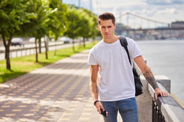 Beyaz tişörtlü ve kot pantolonlu genç bir adam, siyah bir sırt çantası taşıyor, nehir kenarındaki bir patikada korkuluklara yaslanıyor. Arka planda ağaçlar ve bir köprü görünür..