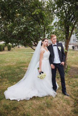 Yeni evliler gün batımında özel bir anı paylaşıyorlar. Gelin beyaz bir elbise ve duvak içinde bir buket beyaz gül tutarken damat nazikçe onu yanında tutuyor..