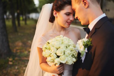 Gelin ve damat gün batımında romantik bir anı paylaşıyorlar. Resmi takım elbiseli damat, geline yakın duruyor. Beyaz bir elbise ve akarsu peçesiyle, altın güneş ışığıyla aydınlatılıyor..