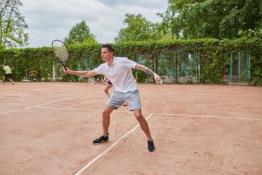 Elinde tenis raketi olan genç bir adam, arka planda bir bayan tenis partneriyle servis ya da dönüş vuruşuna hazırlanıyor..