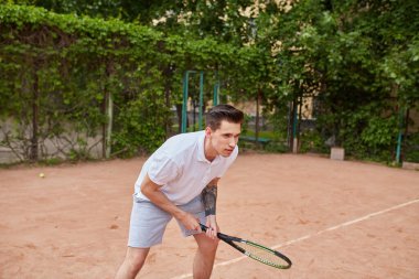 Odaklanmış genç bir adam kil tenis kortunda servis yapmaya hazırlanıyor. Beyaz polo tişört ve gri şort giymiş, elinde bir tenis raketi tutmakta ve harekete geçmeye hazırdır..