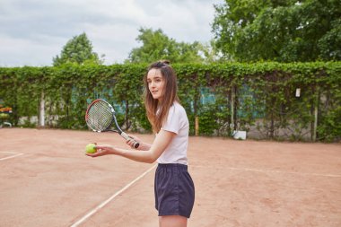 Genç bir bayan tenisçi kil sahada kararlılıkla hazırlanıyor. Beyaz polo ve donanma şortu giymiş, raketini sıkı sıkı tutuyor, yemyeşil bir ortamda harekete geçmeye hazır..
