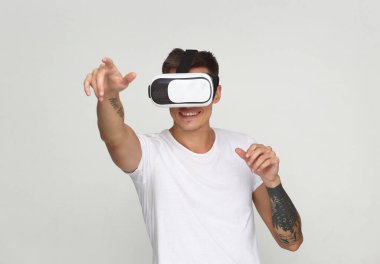 Beyaz tişörtlü genç bir adam VR kulaklık takıyor, sanal bir çevreyle iletişim kuruyor, ellerini kaldırmış, beyaz bir arka planda izole edilmiş..