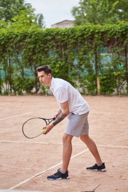 Odaklanmış genç bir adam kil tenis kortunda servis yapmaya hazırlanıyor. Beyaz polo tişört ve gri şort giymiş, elinde bir tenis raketi tutmakta ve harekete geçmeye hazırdır..