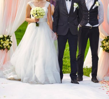 Gelinin elinde beyaz bir gül buketi, damadın klasik bir takım elbiseyle beyaz bir halının üzerinde çiçeklerle süslenmiş bir kemerin altında duran bir sağdıcı gösteren zarif bir düğün sahnesi..