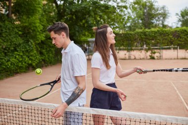 Bir erkek ve bir kadın tenis kortunda sırt sırta, raketleri güvenle tutuyorlar. Takım çalışmasını, odaklanmayı ve kararlılığı iletiyorlar, maç ya da antrenman oturumuna hazırlar..