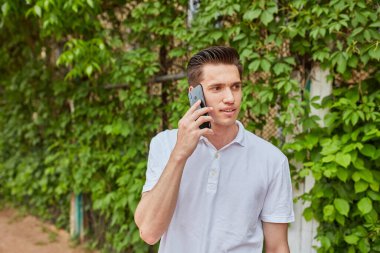 Dışarıda duran genç bir adam kulağına akıllı telefon dayayıp yeşil sarmaşık arka planında beyaz polo tişört giyiyor. Sahne, odak ve sıradan bir atmosfer taşır..